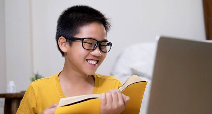 A young boy wearing glasses and a yellow shirt holds a book while looking at his computer screen